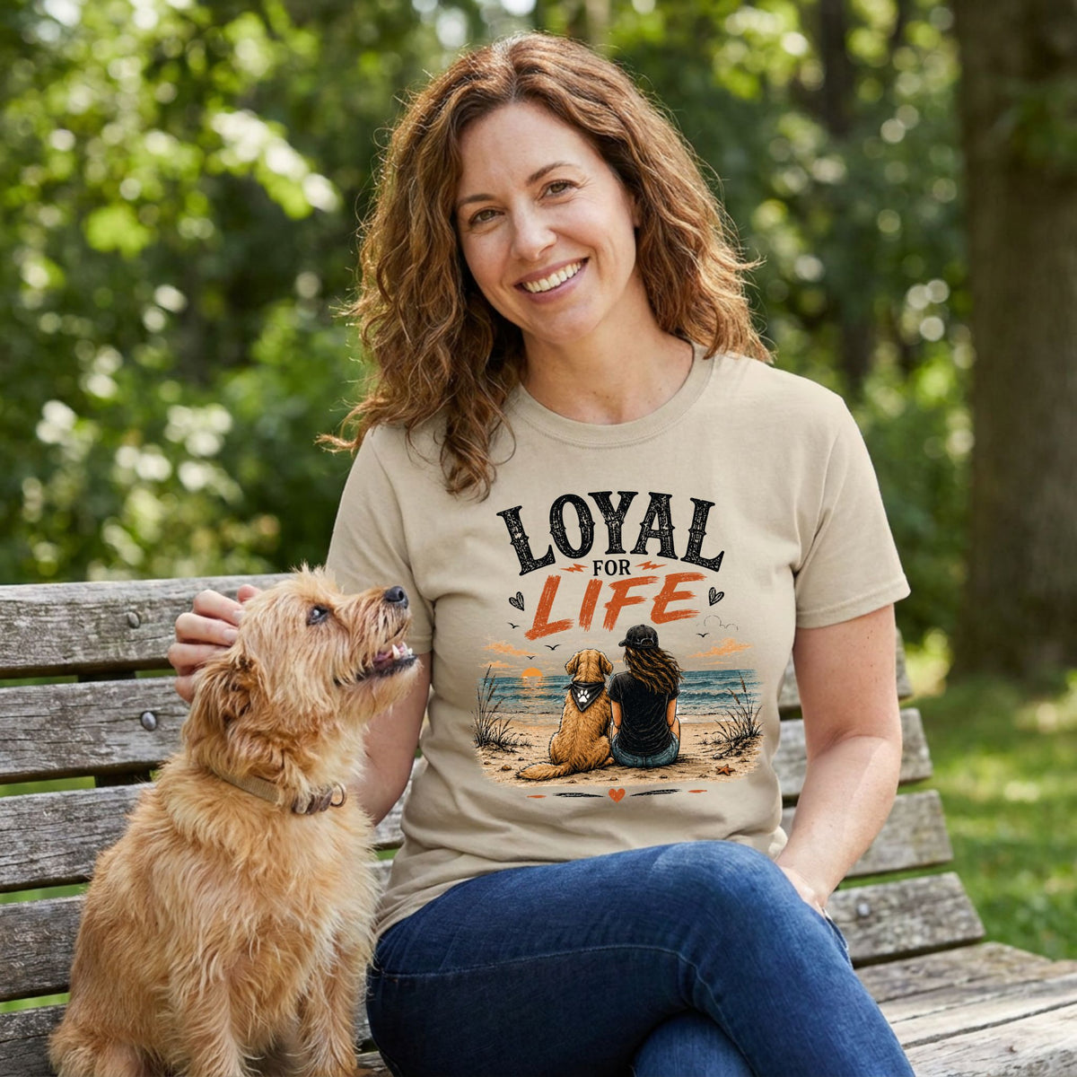 Woman wearing a beige crewneck t-shirt with a Loyal For Life beach graphic, sitting on a wooden bench with her small dog.