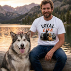 Man wearing a white crew neck t-shirt with a Loyal For Life mountain and dog graphic, sitting by a lake with his dog.