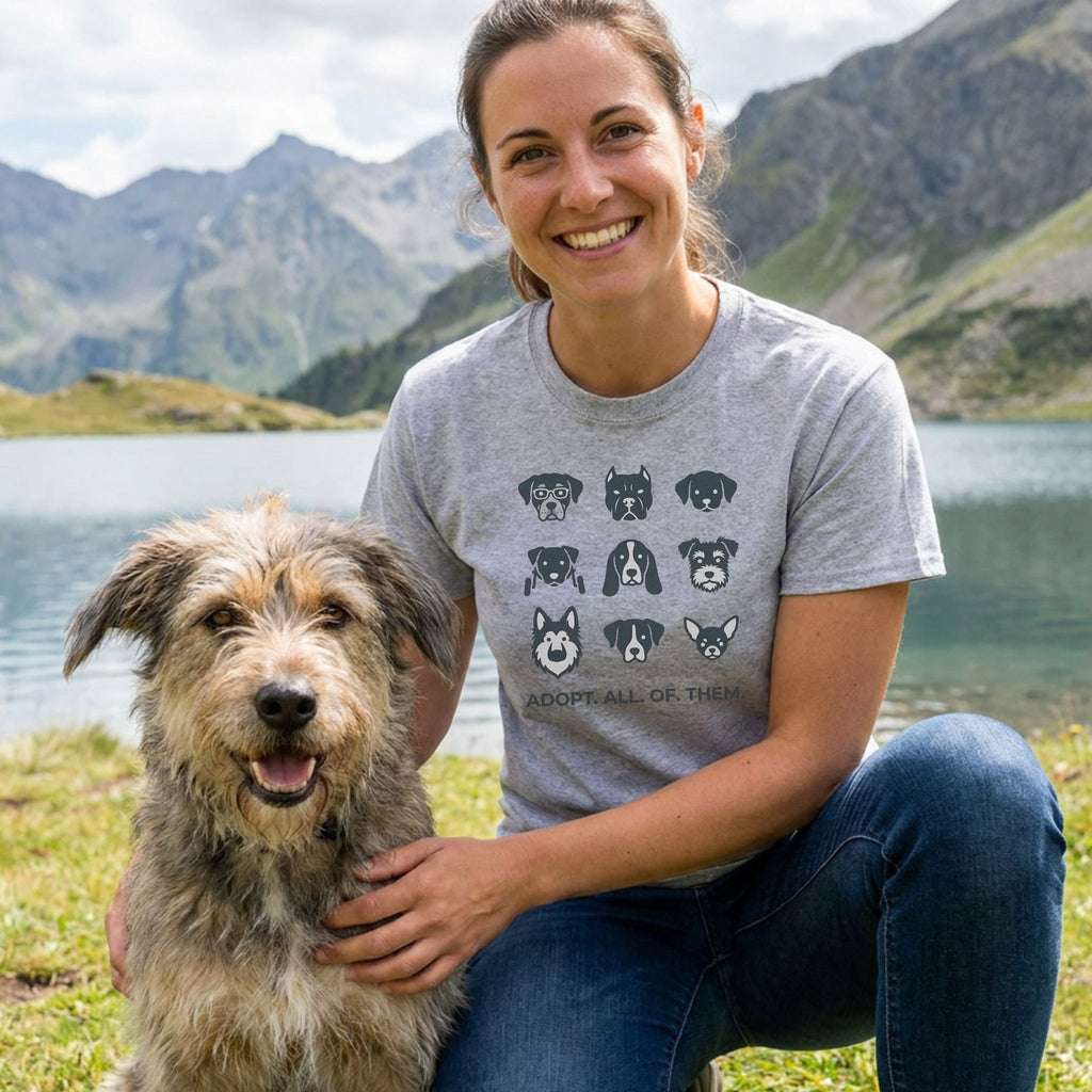 Woman in a grey "Adopt All of Them" t-shirt with dog illustrations, kneeling beside a shaggy dog outdoors.