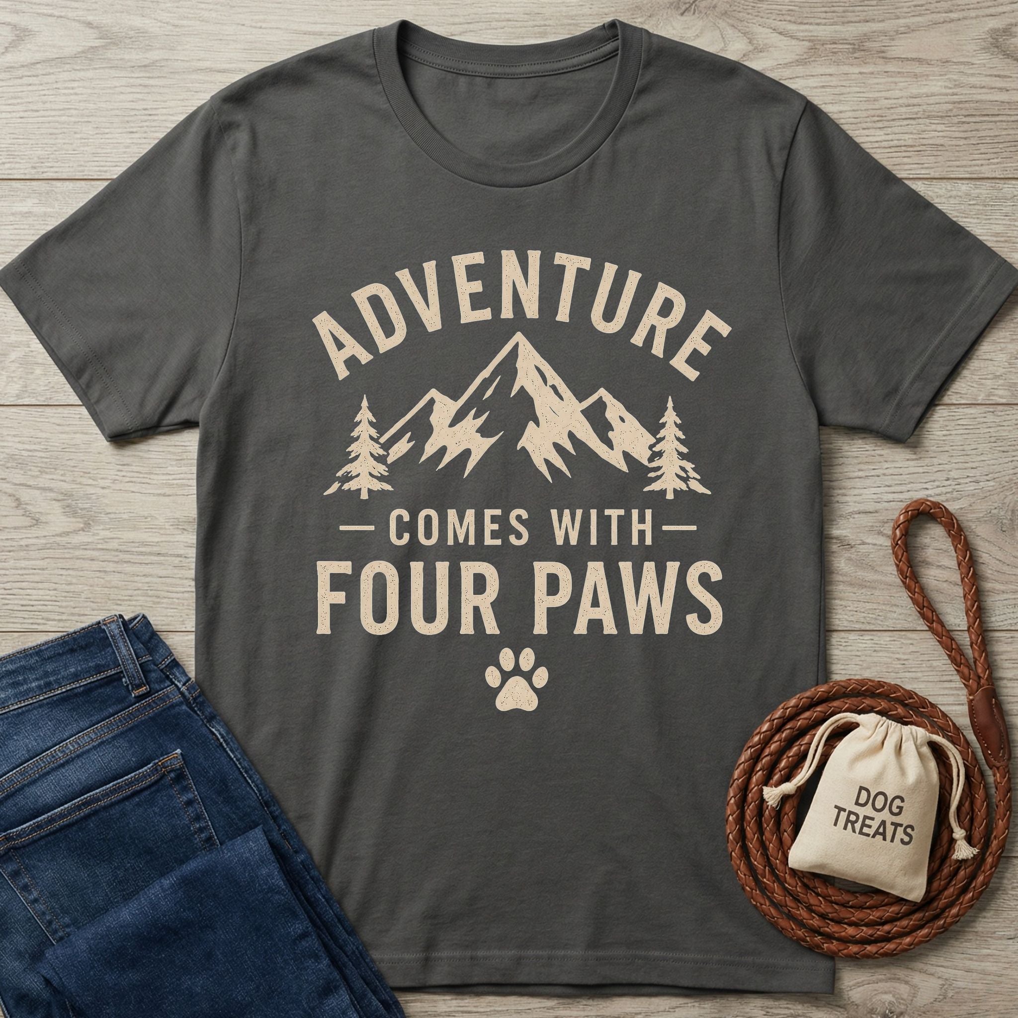 Charcoal gray "Adventure Comes With Four Paws" t-shirt with mountain graphic, next to jeans and dog treats.