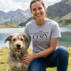 Woman in a gray "Architecture of Cozy" t-shirt with a rescue dog by a lake and mountains.