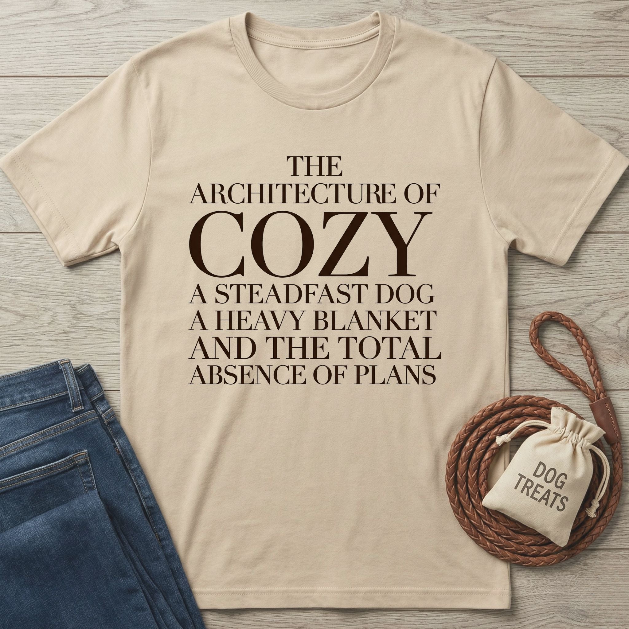 Beige Gildan Softstyle® t-shirt with "The Architecture of Cozy" text, next to jeans and dog treats.