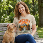 Woman wearing a beige crewneck t-shirt with a mountain, river, and dog graphic, sitting on a bench with her pet dog.