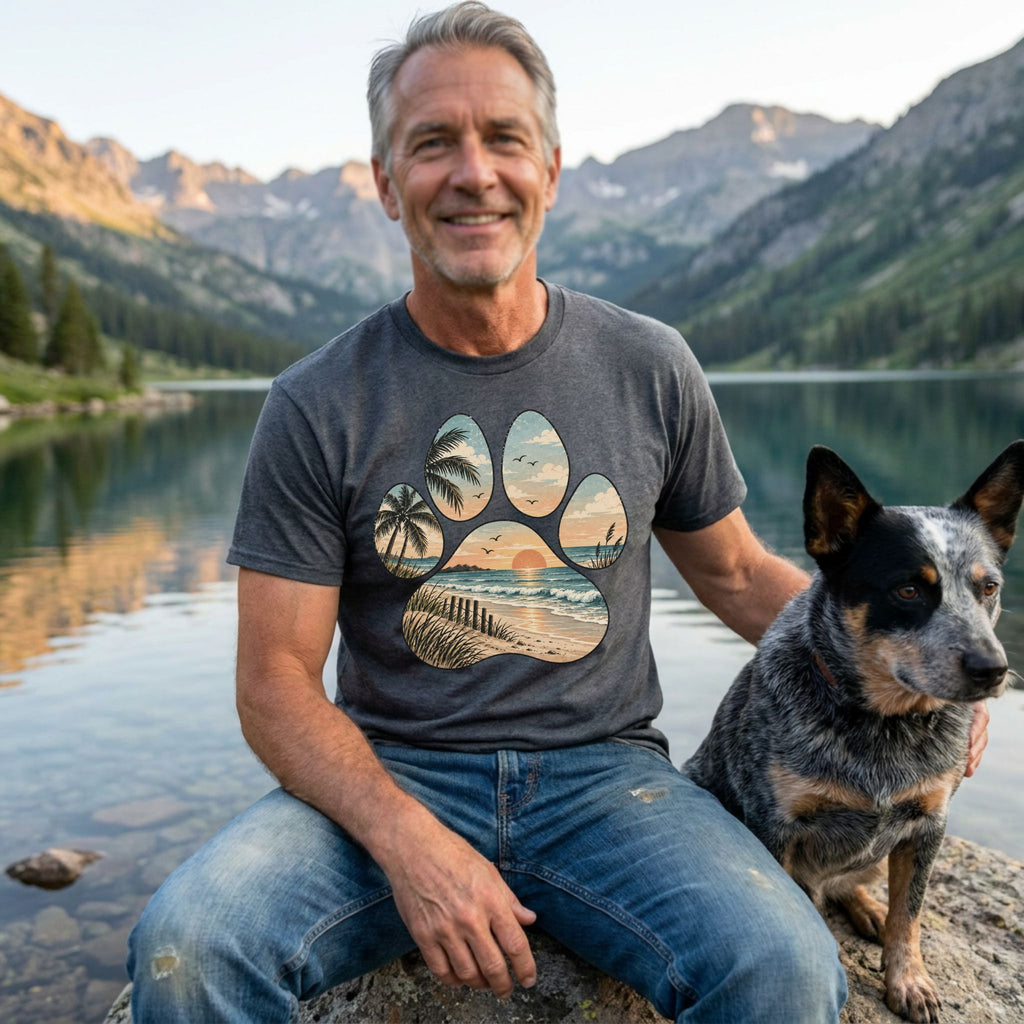 Man wearing a grey t-shirt with a beach sunset and palm tree paw print design while sitting by a lake with his dog.