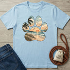 Light blue t-shirt featuring a paw print graphic with a sunset beach, palm trees, and ocean waves design.