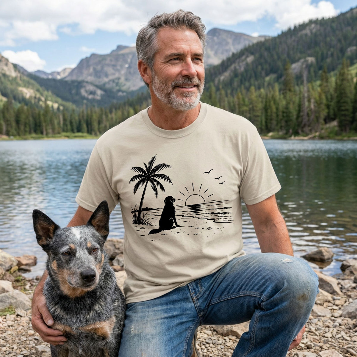 Man wearing a beige crewneck t-shirt featuring a black graphic of a dog, palm tree, and sunset by a mountain lake.