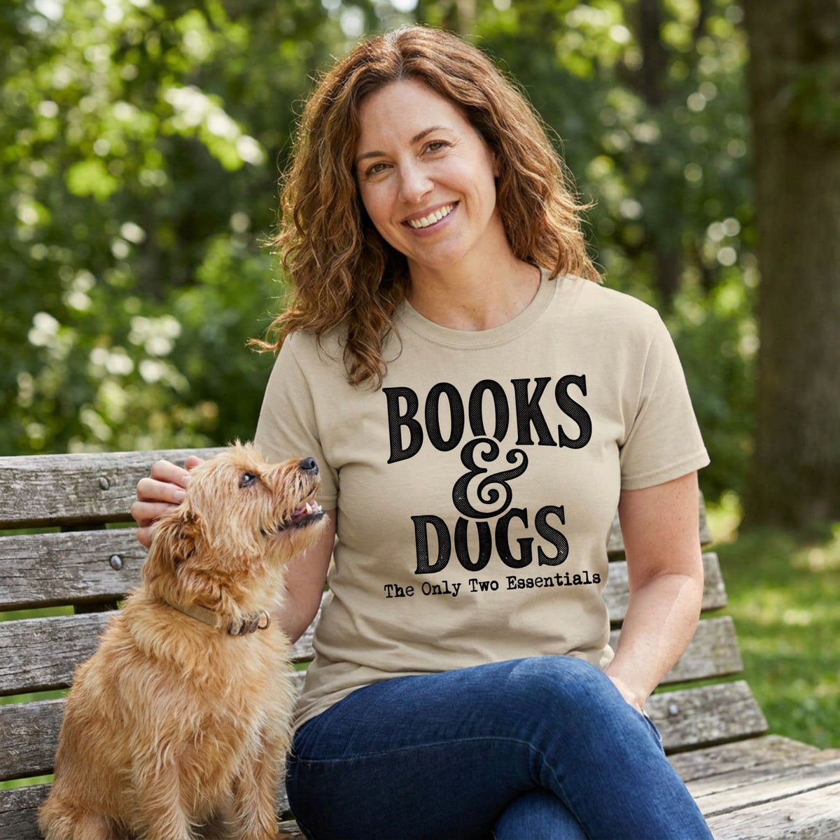 Woman wearing a beige crewneck T-shirt with black Books & Dogs graphic text, sitting on a park bench with a small dog.