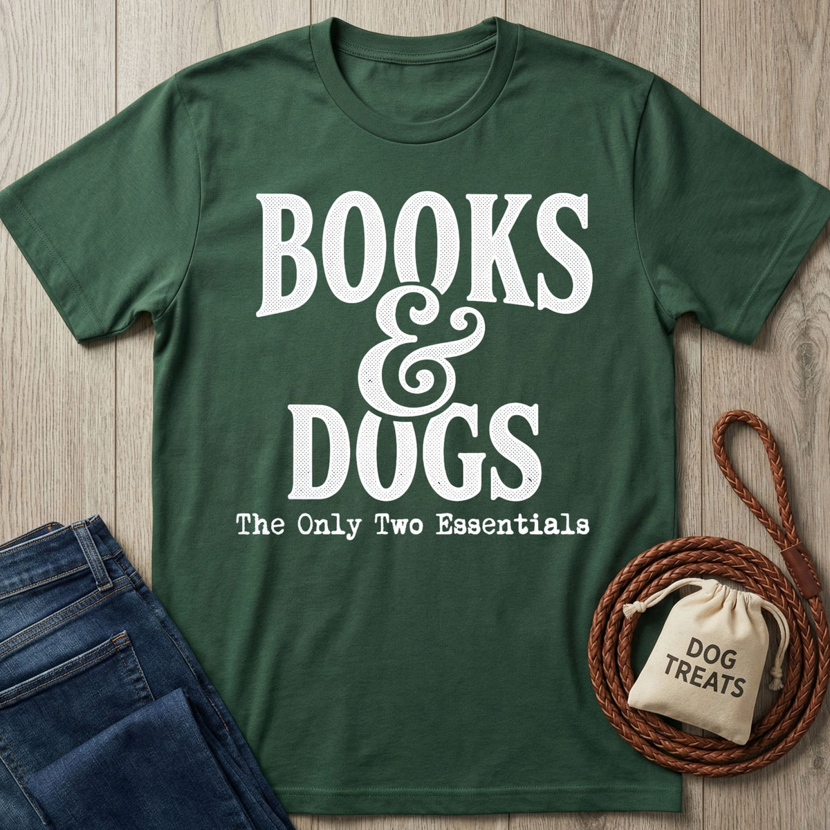 Forest green crewneck T-shirt with white Books & Dogs graphic text, laid flat next to blue jeans and a dog treat bag.