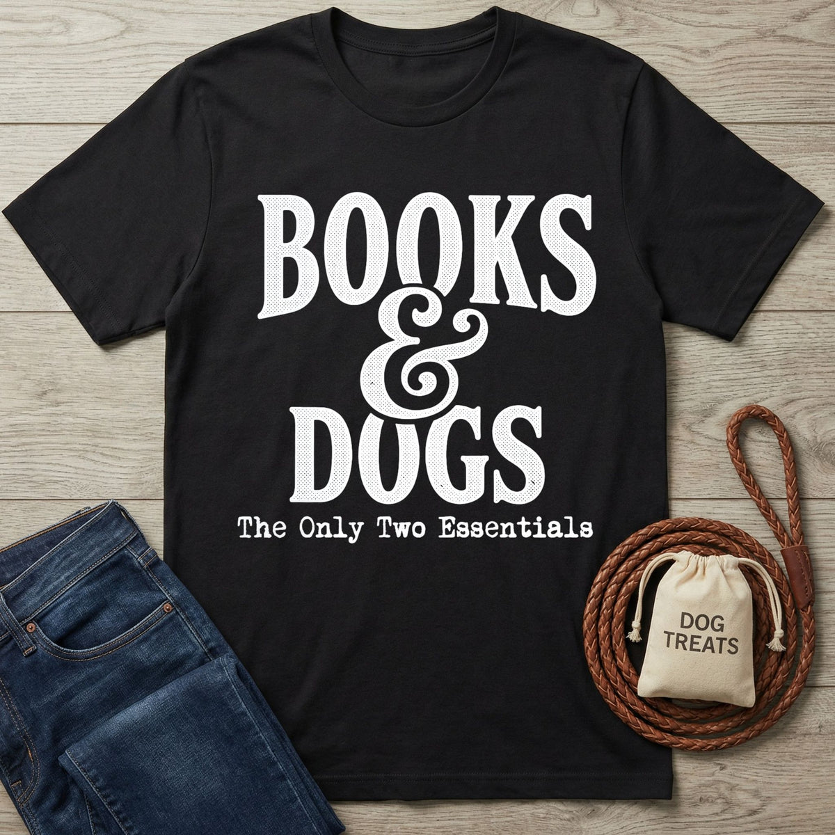 Black crewneck T-shirt with white Books & Dogs graphic text, laid flat next to blue jeans, a leather leash, and a treat bag.