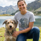 Woman in a grey botanical dog t-shirt with a black lab design, kneeling with a shaggy dog by a lake.