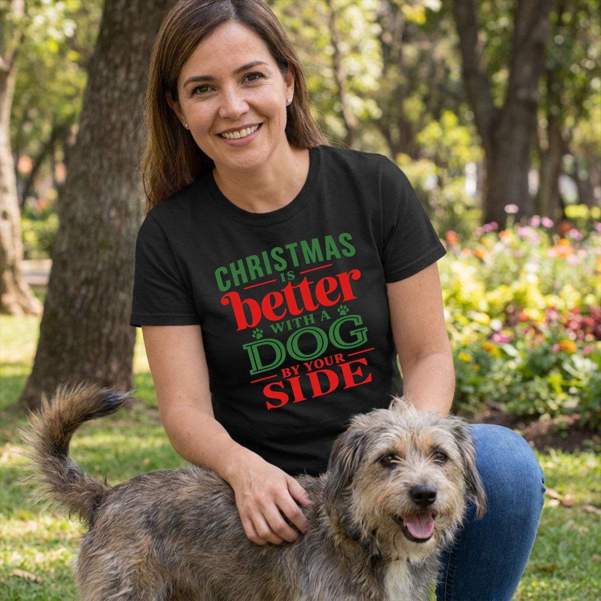 Woman wearing a black crewneck T-shirt with red and green Christmas text, petting her dog in a park.