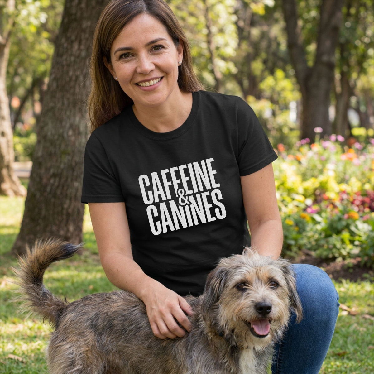 Woman in black "Caffeine & Canines" tee petting a scruffy dog in a park.
