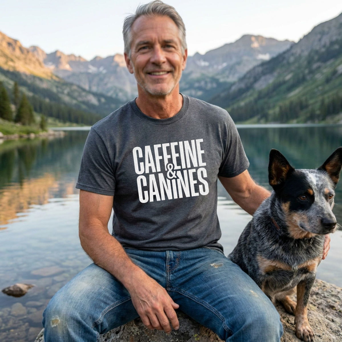 Man in a charcoal grey "Caffeine & Canines" Gildan Softstyle tee with a blue heeler dog by a lake.