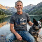Man wearing a charcoal grey crewneck t-shirt featuring a graphic of a dog, campfire, and mountains by a lake.