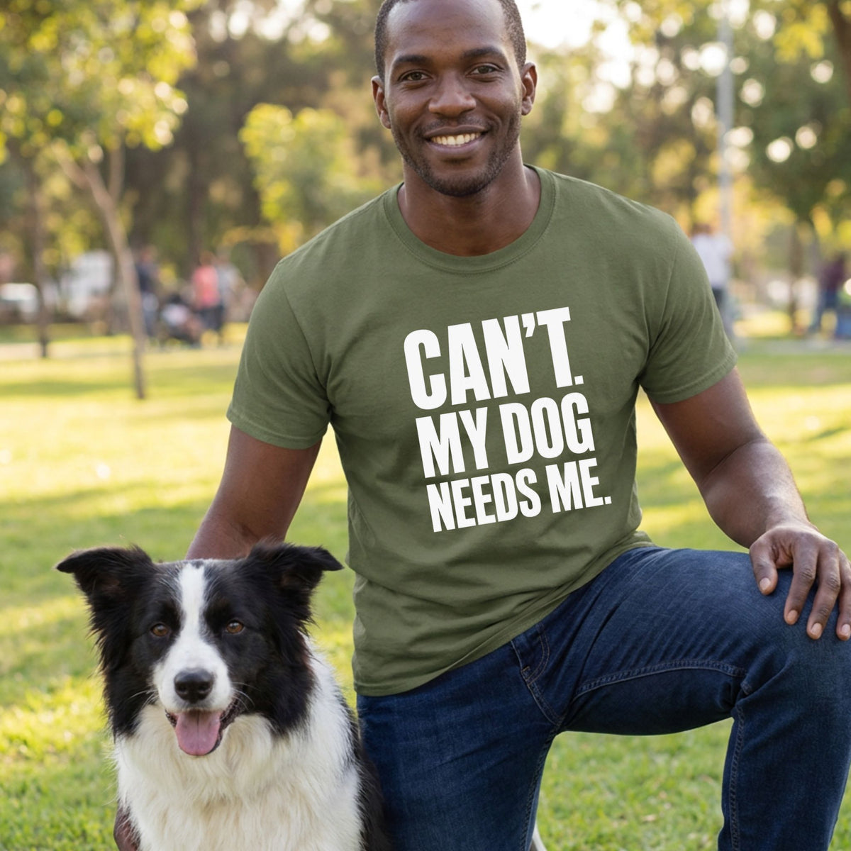 Man wearing an olive green crew neck t-shirt with white text reading Can't. My Dog Needs Me. while kneeling with a dog.