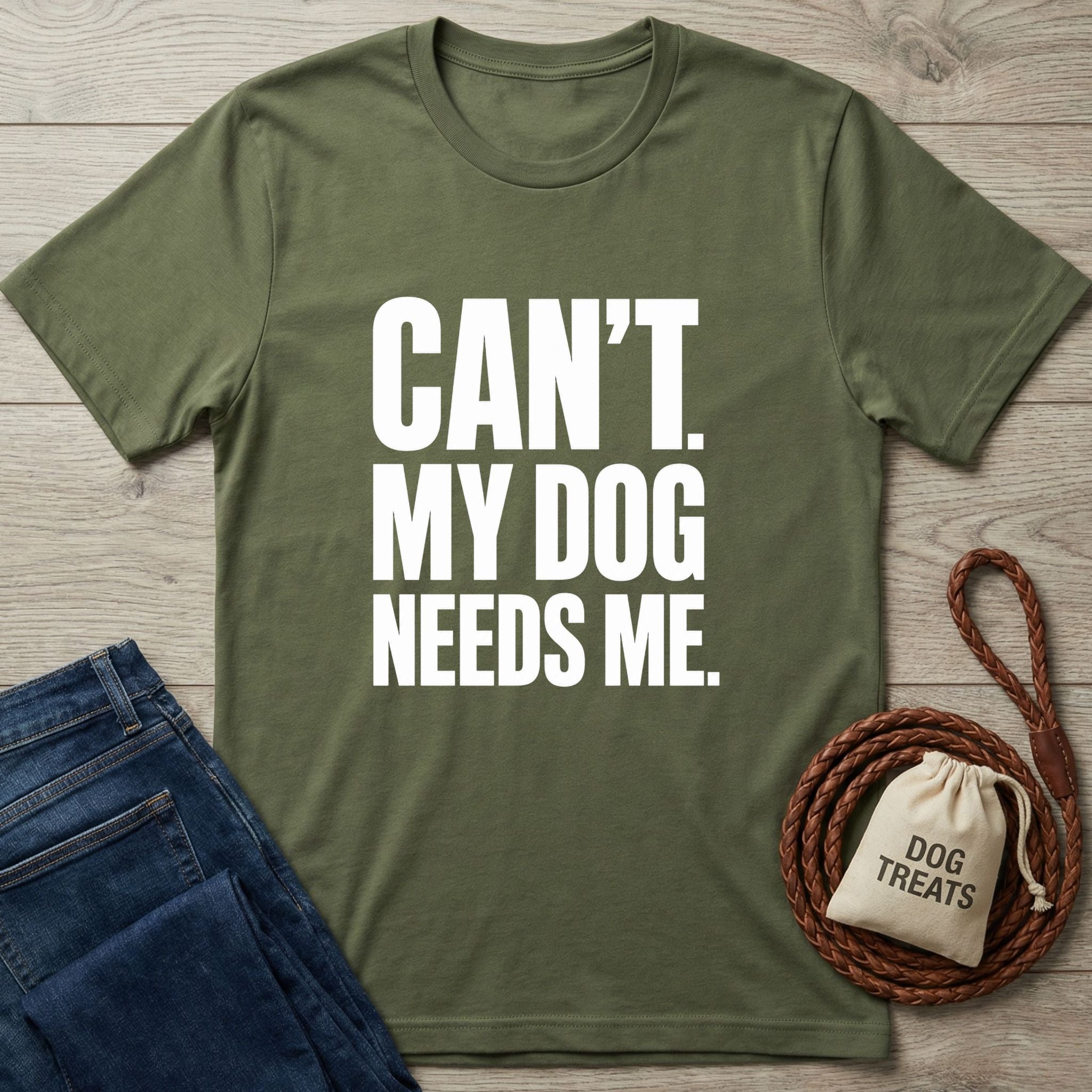 Olive green crewneck t-shirt with white text reading Can't. My Dog Needs Me, laid on wood with jeans and a dog treat bag.