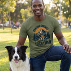 Man wearing an olive green crewneck t-shirt with a mountain, hiker, and dog graphic that reads Chasing Summits Trails Are Better Together.