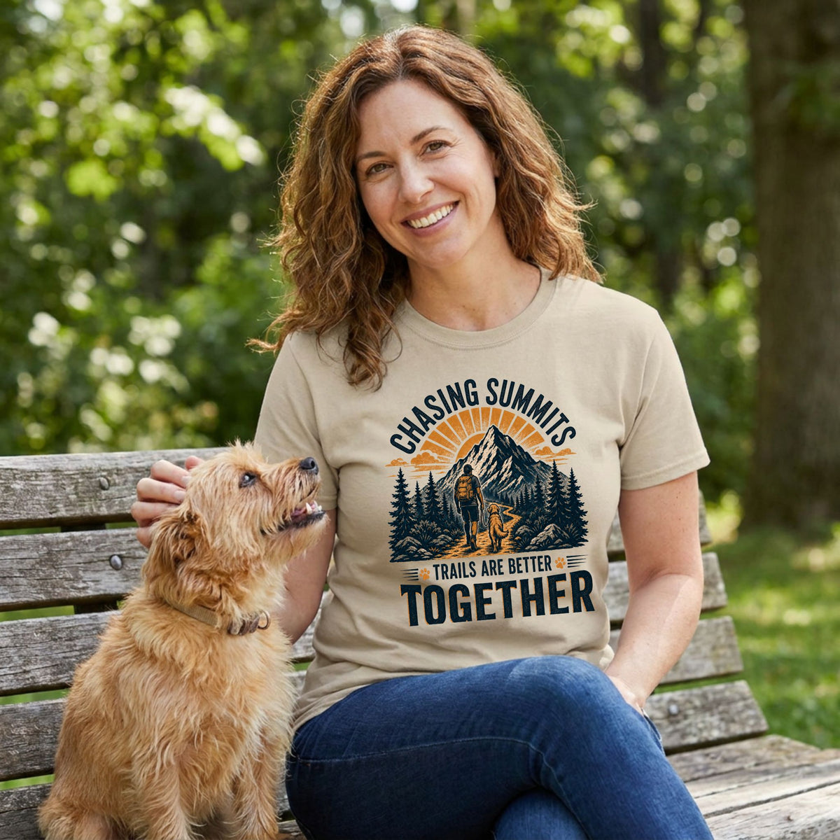 Woman wearing a tan crewneck t-shirt featuring a mountain hiking graphic with a person and dog, sitting on a park bench.