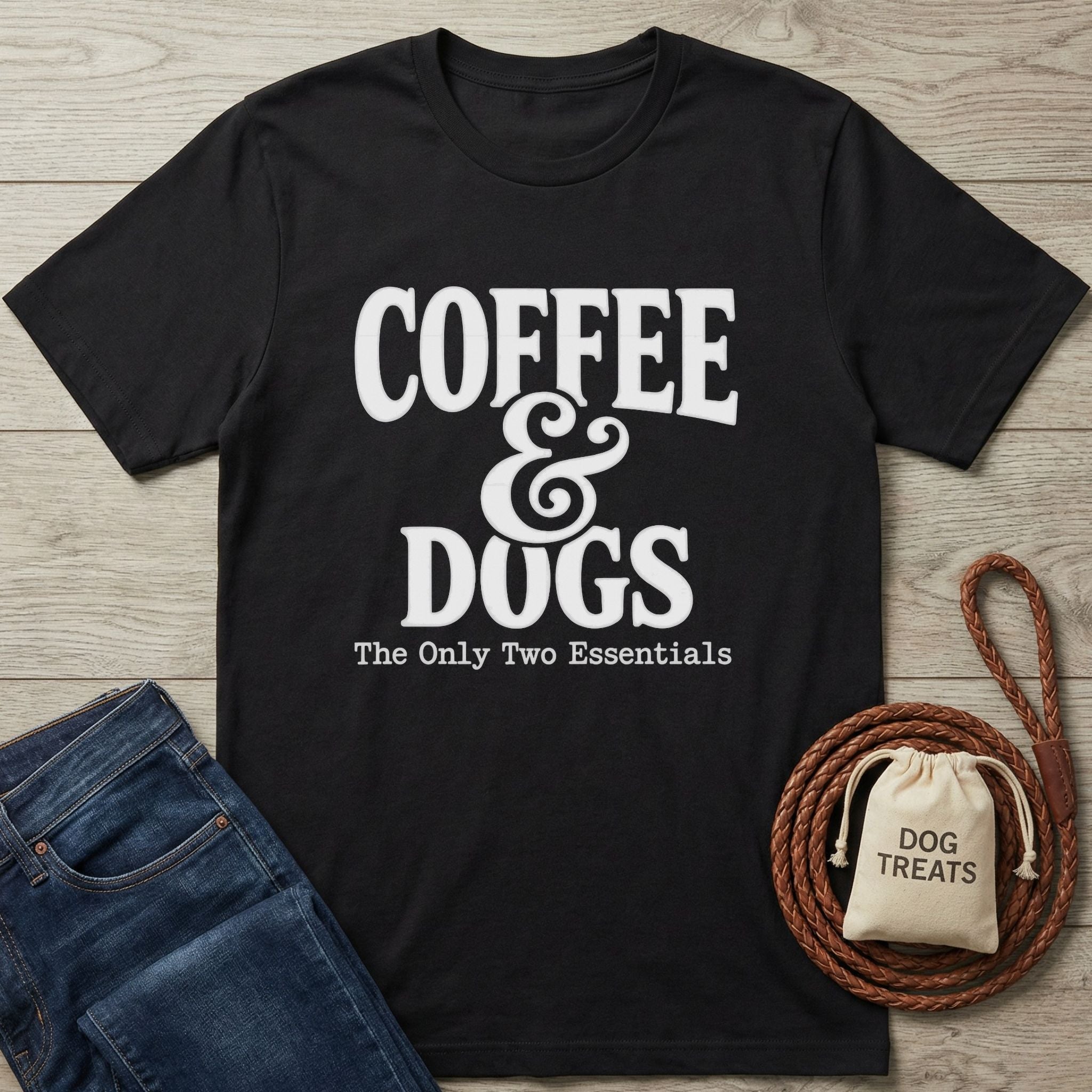 Black Gildan Softstyle® t-shirt with "Coffee & Dogs: The Only Two Essentials" text, next to jeans and dog treats.