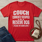 Red Gildan Softstyle® t-shirt with "THE COUCH IS CURRENTLY OCCUPIED BY A RESCUE DOG AND HIS HUMAN PLEASE TRY AGAIN LATER" in white text, beside jeans and dog treats.