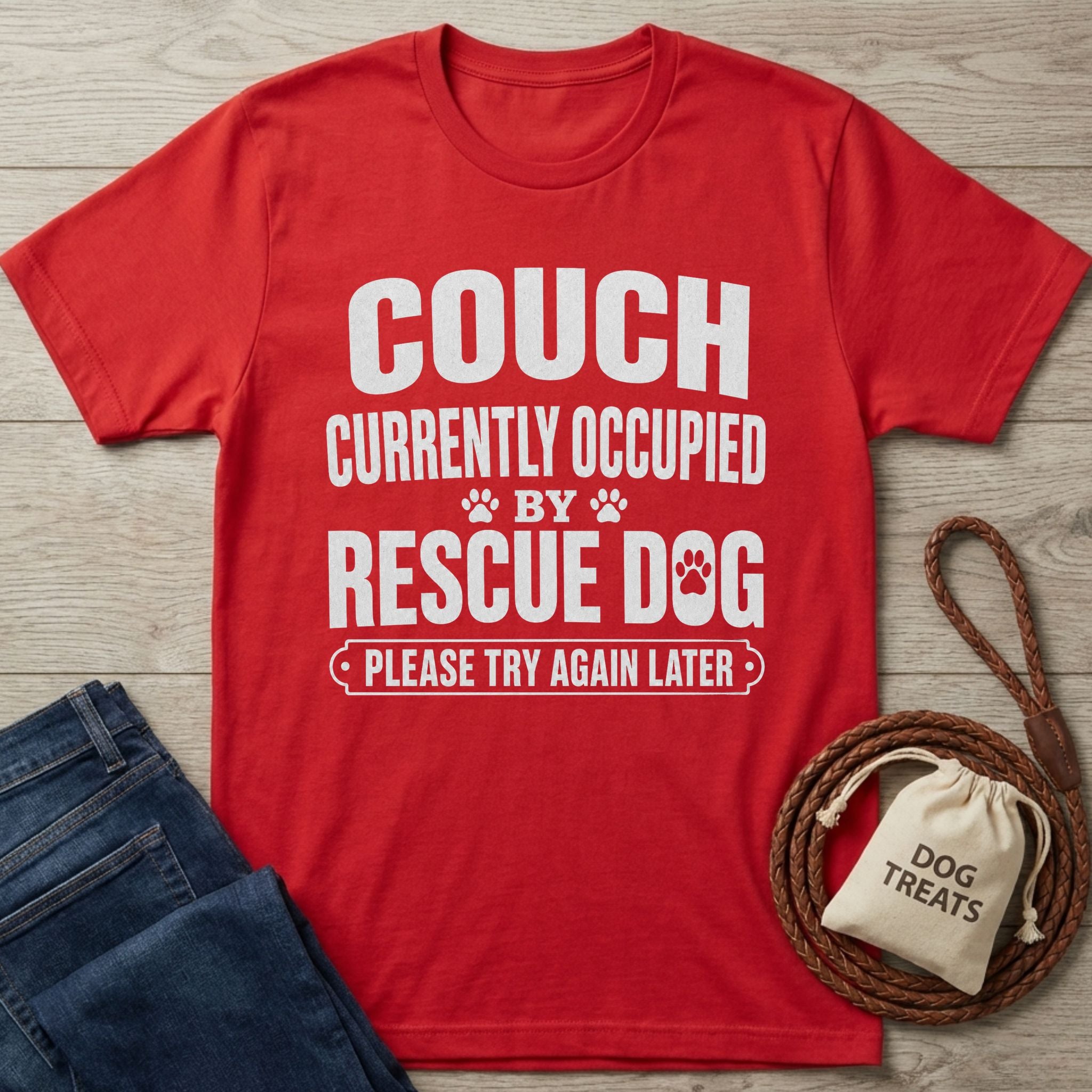 Red Gildan Softstyle® t-shirt with "THE COUCH IS CURRENTLY OCCUPIED BY A RESCUE DOG AND HIS HUMAN PLEASE TRY AGAIN LATER" in white text, beside jeans and dog treats.