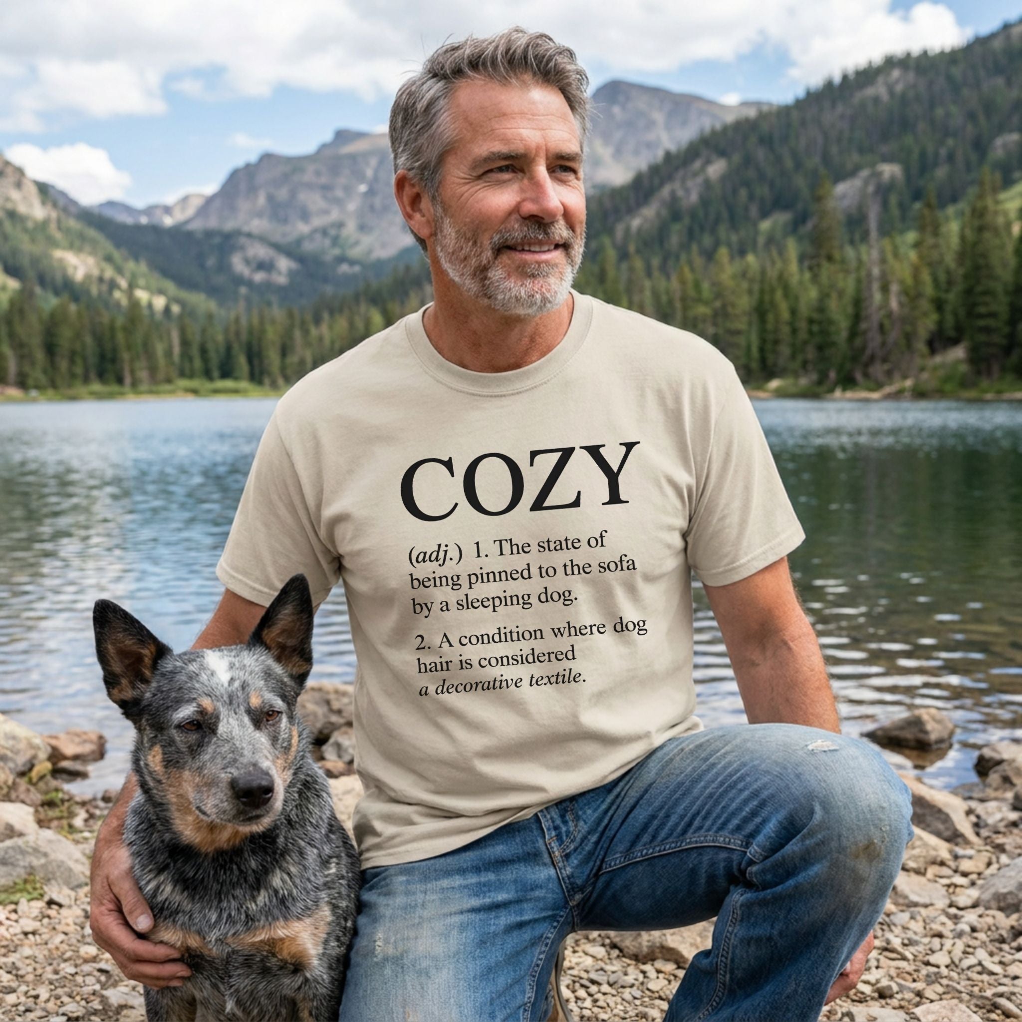 Man in a sand-colored "COZY" definition tee with a blue heeler dog by a mountain lake.