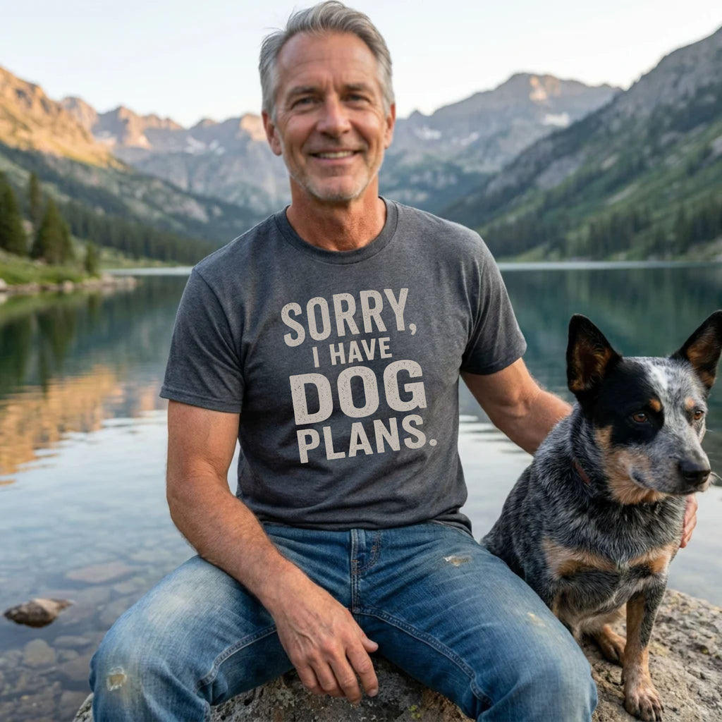 Man wearing a charcoal grey crewneck t-shirt with the white text Sorry, I Have Dog Plans sitting by a lake with his dog.