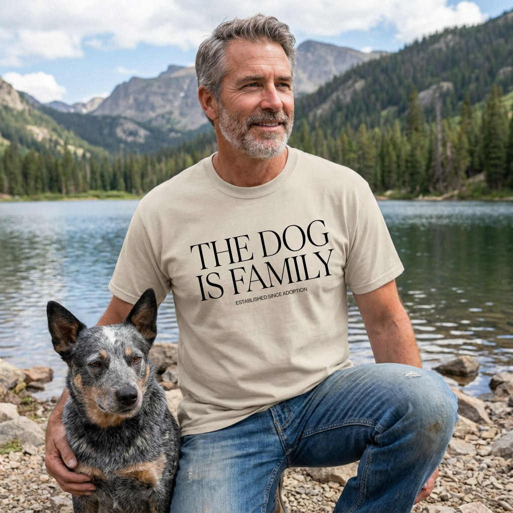 Man in a beige "THE DOG IS FAMILY" Gildan Softstyle® tee with a blue heeler dog by a mountain lake.