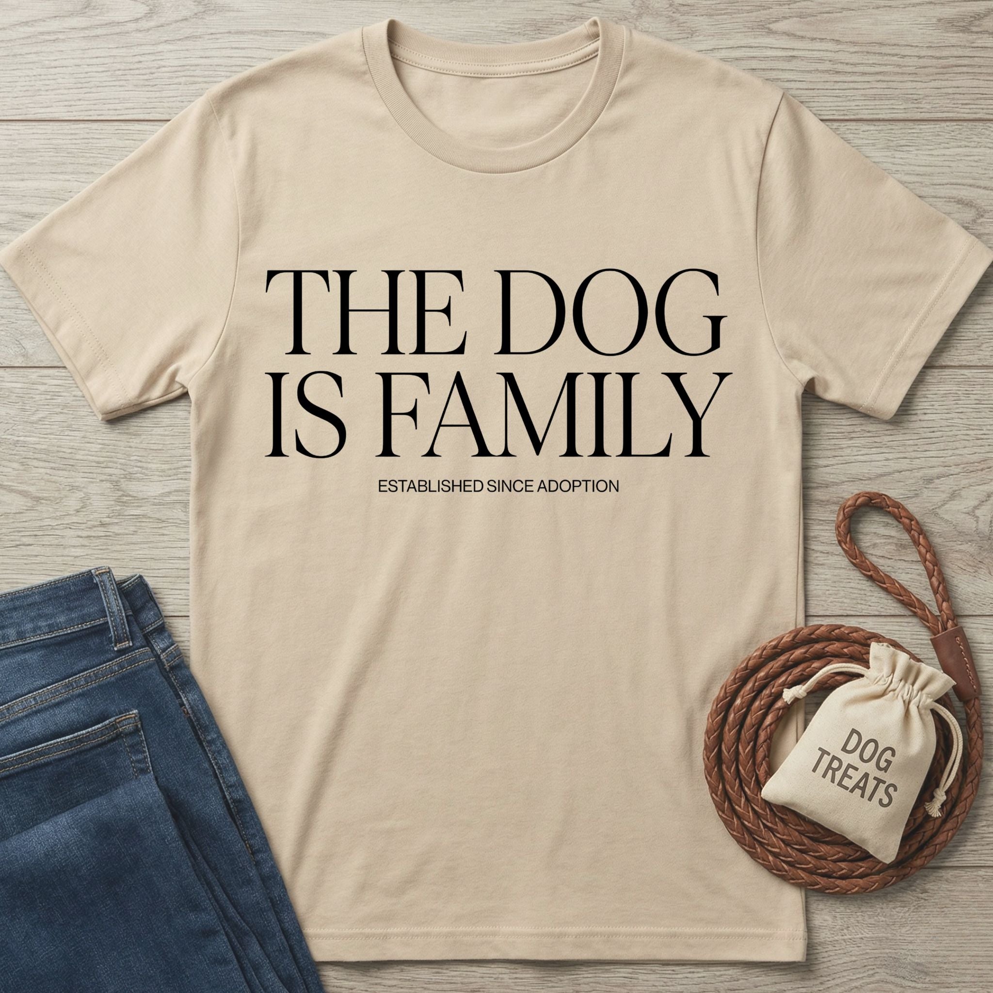 Beige Gildan Softstyle® t-shirt with "THE DOG IS FAMILY" text, next to jeans and dog treats.