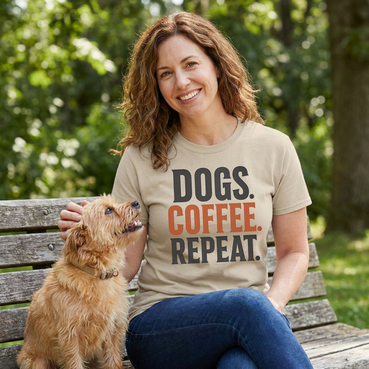 Woman wearing a beige crewneck t-shirt with Dogs, Coffee, Repeat printed in black and orange text while sitting with a dog.