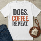 White cotton t-shirt featuring the text Dogs Coffee Repeat in bold black and orange lettering on a wooden background.