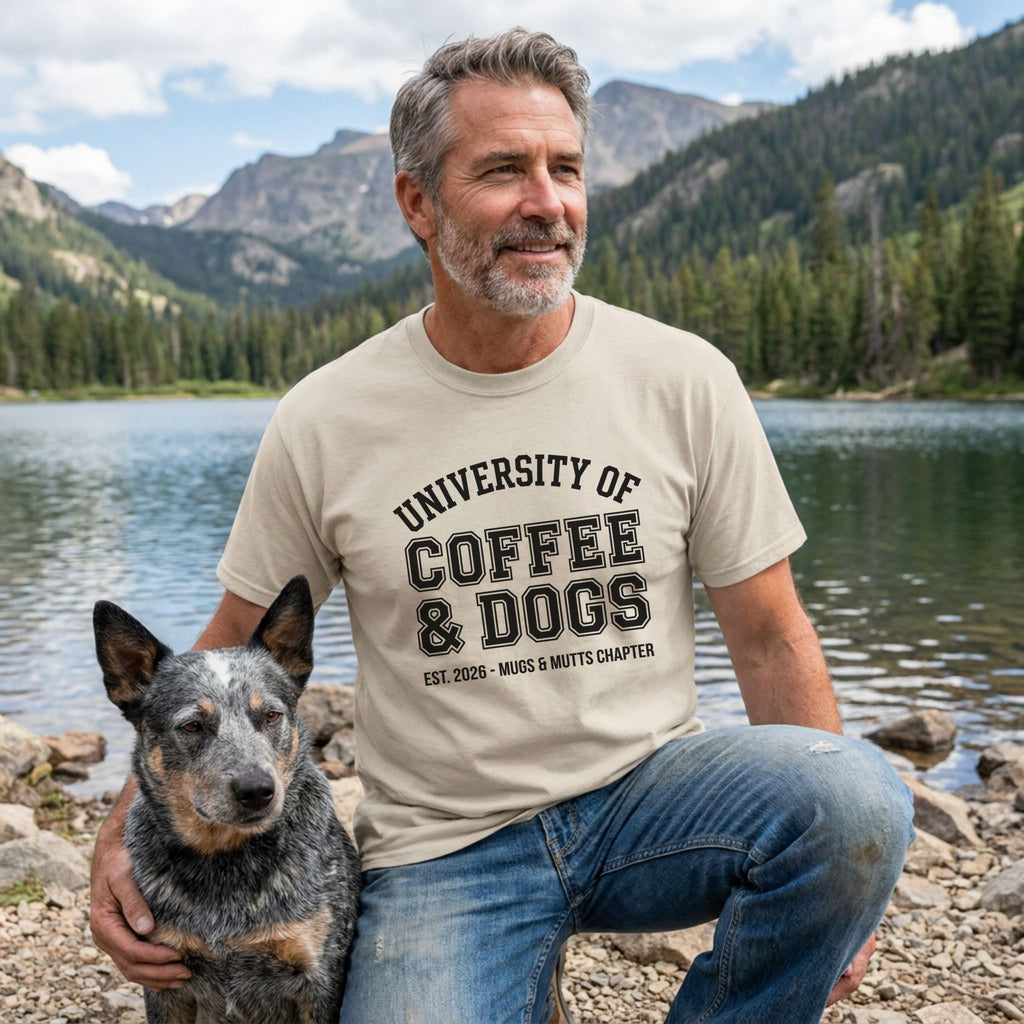 Man in a light beige "University of Coffee & Dogs" t-shirt with a blue heeler dog by a lake.