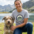 Woman in a grey "The Dogs > People Club" t-shirt with a dog, mountains, and lake in the background.