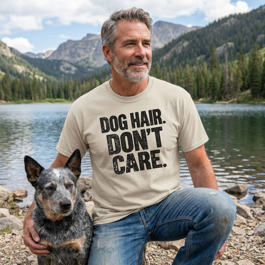 Man wearing a beige crewneck t-shirt with the text DOG HAIR. DON'T CARE. in bold black letters, posing with a dog by a lake.