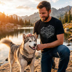 Man in black "Elevation Lines" tee petting a husky by a lake with mountains at sunset.