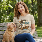 Woman wearing a beige crewneck t-shirt with a graphic of a dog in sunglasses holding a coffee cup, sitting on a park bench.