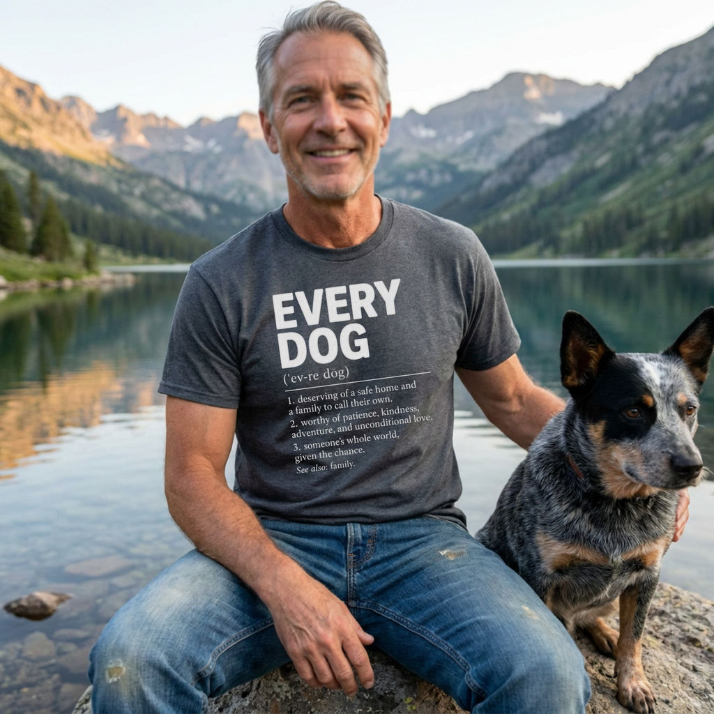 Man wearing a charcoal grey Every Dog definition graphic t-shirt while sitting by a mountain lake with his dog.