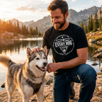 Man wearing a black Every Dog graphic t-shirt while petting a husky by a mountain lake at sunset.