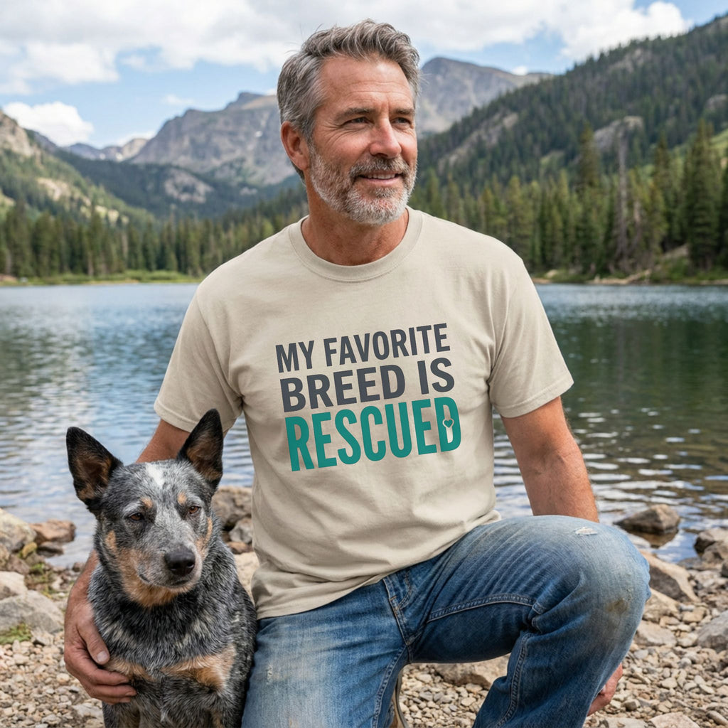 Man in a beige "My Favorite Breed Is Rescued" t-shirt with a blue heeler dog by a lake.