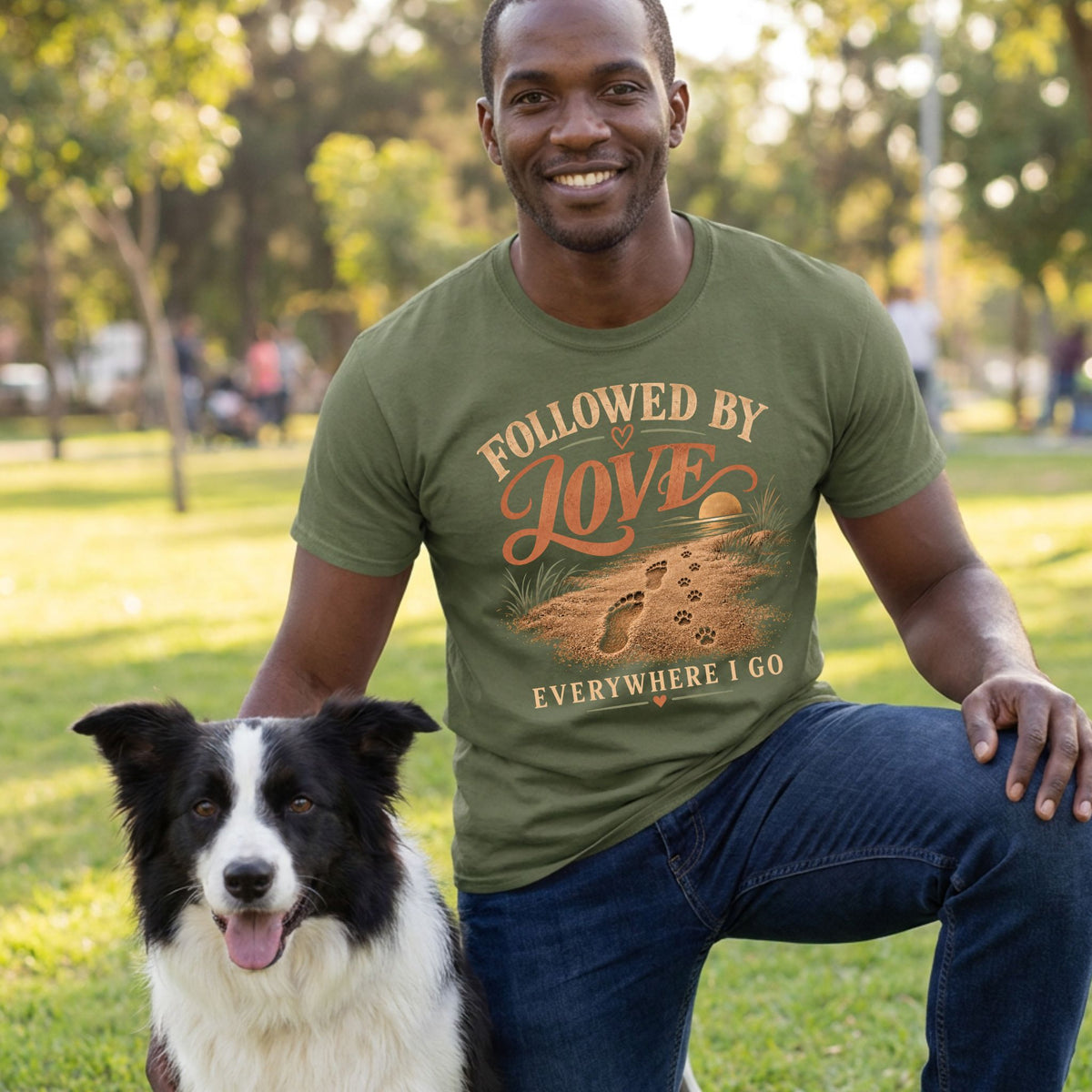 Man wearing an olive green T-shirt with a beach-themed paw print graphic, kneeling outdoors next to a border collie.
