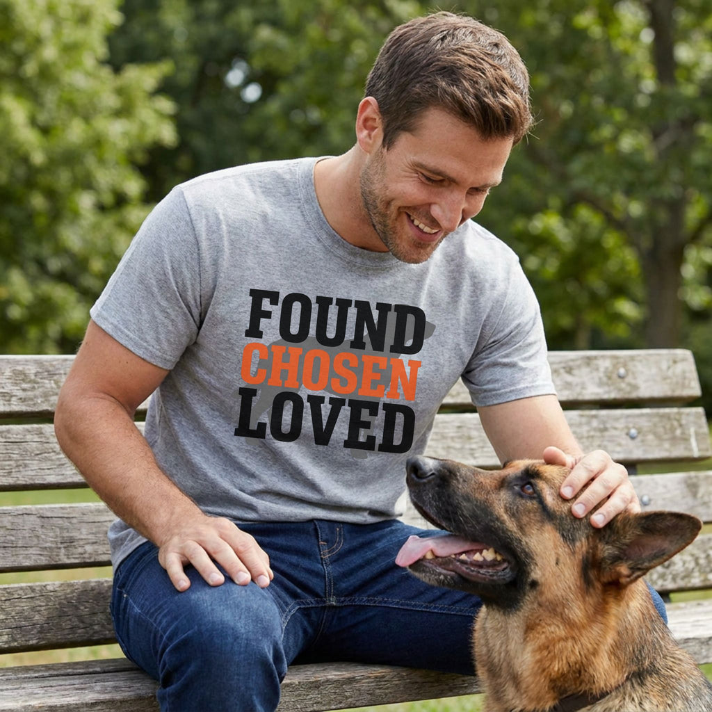 Man in a grey "Found Chosen Loved" t-shirt petting a German Shepherd on a park bench.