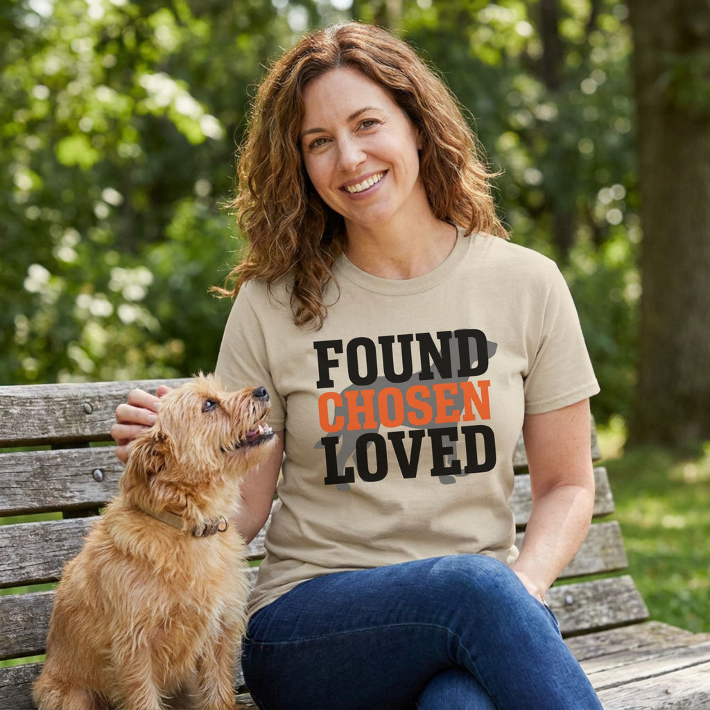 Smiling woman in a tan "Found Chosen Loved" t-shirt petting a small brown dog on a park bench.