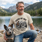 Man in a beige "Found My Way" graphic tee with a hiking dog design, kneeling by a lake with a blue heeler.