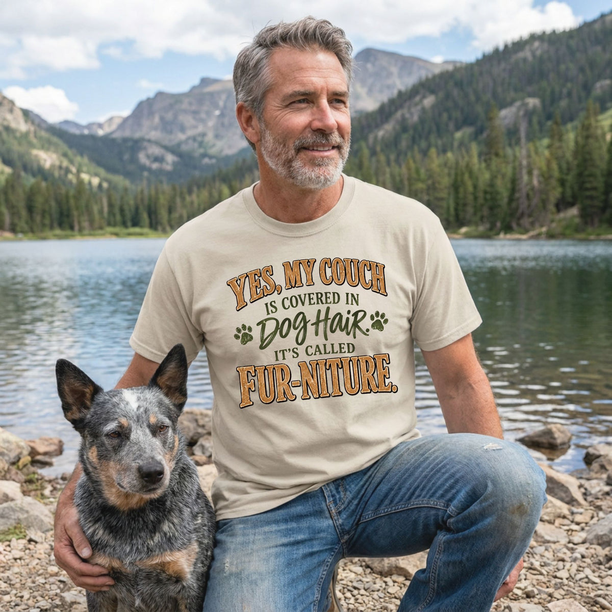 Man wearing a beige graphic T-shirt with the text Fur-niture and paw prints, posing with a dog by a mountain lake.