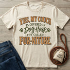 Cream-colored graphic T-shirt with the text Yes, My Couch Is Covered In Dog Hair, It's Called Fur-niture on a wooden background.