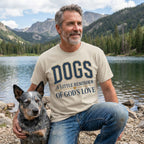 Man wearing a beige crewneck t-shirt with Dogs A Little Reminder Of God's Love text, posing with a dog by a mountain lake.