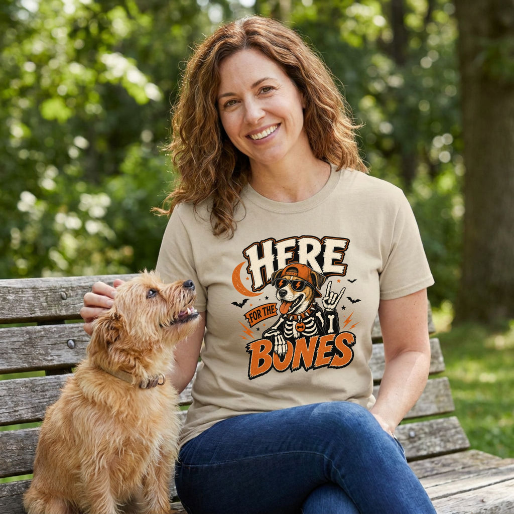 Woman wearing a beige Here For The Bones graphic t-shirt featuring a skeleton dog, sitting on a bench with her terrier.