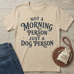 Beige crewneck t-shirt with dark blue "Not a morning person, just a dog person" text, styled with jeans and a dog treat bag.