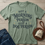 Sage green crew neck t-shirt with Not a Morning Person Just a Dog Person text, laid flat with jeans, a leash, and a treat bag.
