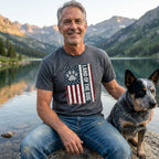 Man wearing a charcoal grey Land of the Dog t-shirt featuring a distressed American flag and paw print design by a lake.